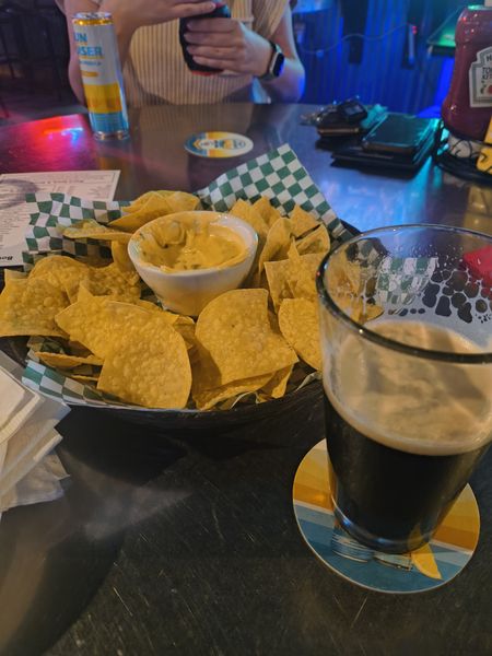The image shows a casual dining setting with a plate of tortilla chips and a bowl of cheese dip on a checkered paper, accompanied by a glass of dark beer on a coaster with a colorful pattern. In the background, a person is holding a can of "Sun Crusher" beer, and a bottle of Heinz ketchup is visible on the counter. The setting appears to be a bar or restaurant with dim lighting and a dark, reflective table surface.