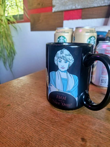 A black ceramic mug featuring a stylized illustration of a white-haired woman in a blue top and the text "NO, I WILL not HAVE A Nice Day!" sits on a wooden table. In the blurred background, cream-colored Starbucks cans and a white Diet Coke can are visible against a wall with wood and red paneling.
