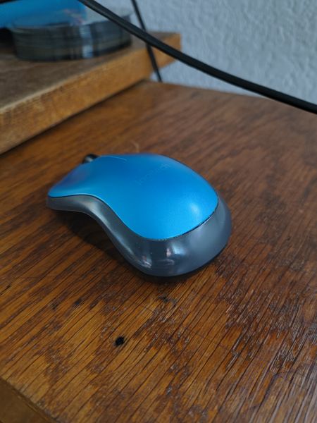 The image features a Logitech Ergo M575 wireless mouse with a blue top and gray bottom, resting on a wooden desk. The background includes a wooden shelf with a glass container and a textured white wall.