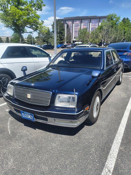 The image features a dark blue Toyota Century sedan parked in a sunny parking lot. The car's license plate reads "ELEGNCY," and it has a distinctive chrome grille and white-walled tires. The setting includes other parked cars and a modern building with large windows in the background, under a clear blue sky.