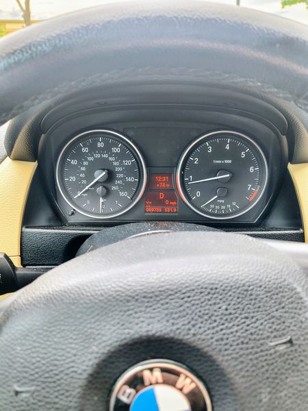 The image shows the dashboard of a BMW vehicle, featuring a black steering wheel with a BMW logo at the center. The dashboard displays two circular gauges: the left gauge is for speed, marked up to 160 mph, and the right gauge is for engine RPM, marked up to 6,000 RPM. The digital display in the center shows the time as 12:31 and the temperature as +74°F. The car's odometer reads 069735 miles. The interior has a black and yellow color scheme.