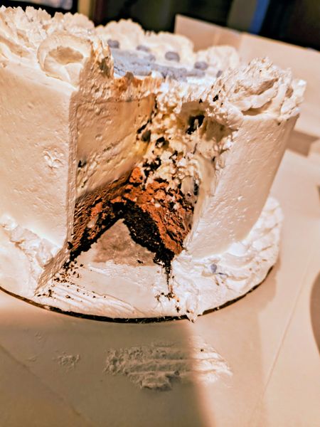 The image shows a partially eaten chocolate cake with white frosting, placed on a white plate. The cake has a rich, dark chocolate interior and is topped with a generous layer of white frosting that appears slightly textured. The setting is indoors, and the background is blurred, focusing attention on the cake.