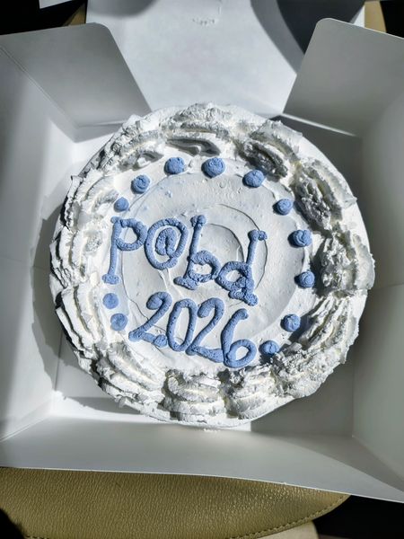 The image shows a round cake with white frosting and blue icing decorations. The cake is placed in a white cardboard box, and the icing spells out "P@bq" and "2026" in a playful, cursive style. The setting appears to be indoors, with a glimpse of a light-colored surface beneath the box.