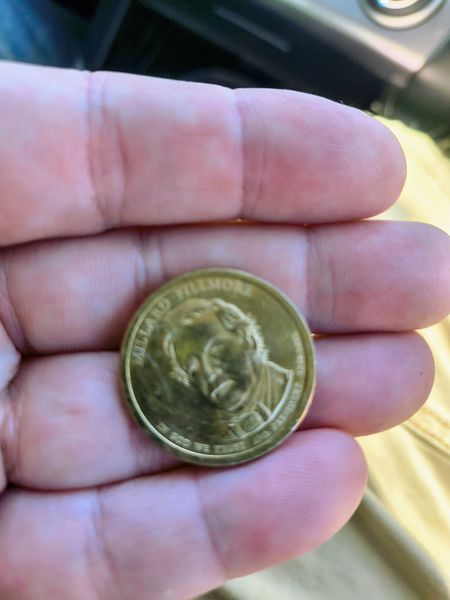 The image shows a hand holding a gold-colored coin with the inscription "Millard Fillmore" on its edge, featuring a detailed portrait in the center. The setting appears to be outdoors, with a blurred background suggesting a vehicle interior and sunlight.
