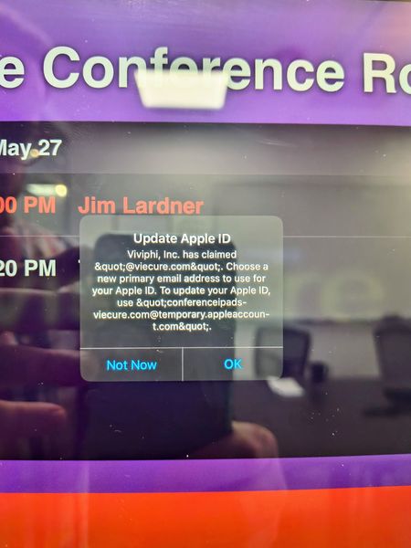 The image shows a computer screen displaying a conference room schedule with a pop-up message from Apple requesting an update to the Apple ID. The message mentions Viviphii, Inc. and provides a temporary email address for the Apple ID update. The screen background includes a purple header with "Conference Room" text and a partially visible date and time for a meeting with Jim Lardner. The bottom of the screen has a red bar.