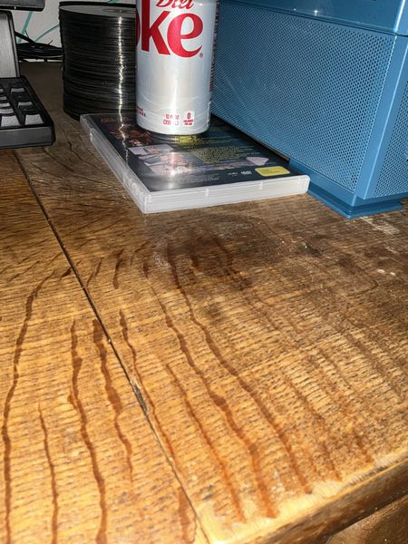 The image shows a close-up of a wooden desk with a Diet Coke can placed on top of a DVD case. The desk has visible wood grain and a natural finish. A blue speaker is partially visible on the right, and a stack of CDs or DVDs is seen in the background.