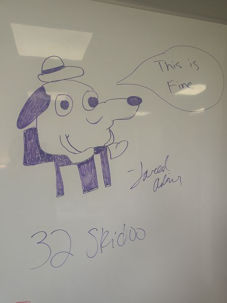 The image shows a whiteboard with a drawing of a dog wearing a hat and sitting on a chair, with a speech bubble saying "This is Fine." The drawing is done in purple marker. Below the drawing, the text "32 Skidoo" is written, and the artist's signature, "Jared Adams," is also visible. The setting appears to be an indoor space, possibly a classroom or meeting room, with a ceiling light reflecting on the whiteboard.
