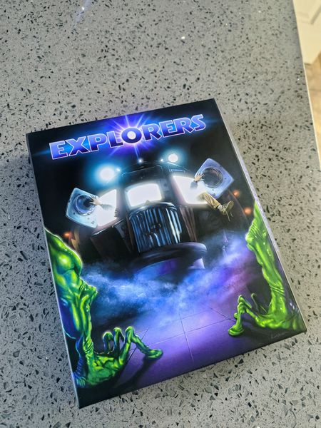 The image features a book titled "Explorers" resting on a speckled gray countertop. The book cover showcases a vintage-style truck with large headlights, emitting a bright glow, and is surrounded by eerie green alien-like hands reaching out from the bottom corners. The title "Explorers" is prominently displayed in a bold, futuristic font with a blue and purple gradient background.
