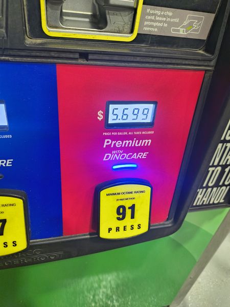 The image shows a gas pump displaying a price of $5.699 per gallon for premium gasoline with Dinocare, branded in white text on a red background. A yellow sticker indicates a minimum octane rating of 91, with the word "PRESS" below it. The pump has a black and yellow design, with a digital display and a slot for a chip card.