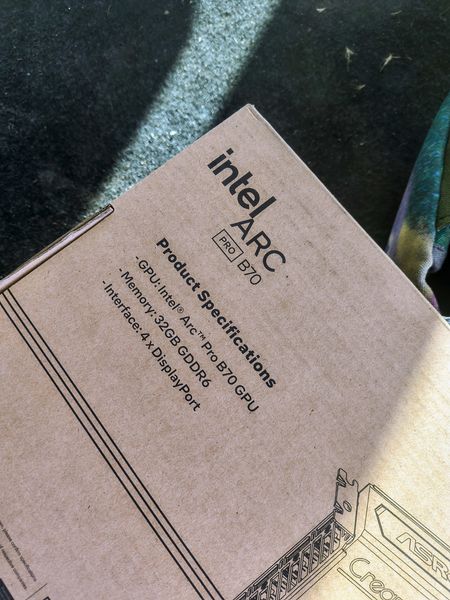 The image shows a brown cardboard box for the Intel ARC Pro B70 graphics card, placed on a textured surface. The box lists specifications including 32GB GDDR6 memory and 4 x DisplayPort interfaces, with the Intel logo and model name prominently displayed.