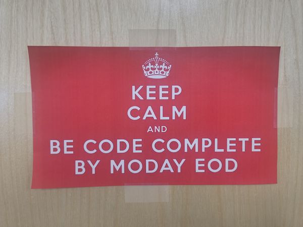 The image features a red poster with white text that reads "KEEP CALM AND BE CODE COMPLETE BY MODAY EOD," accompanied by a crown symbol at the top. The poster is taped to a light wooden surface.