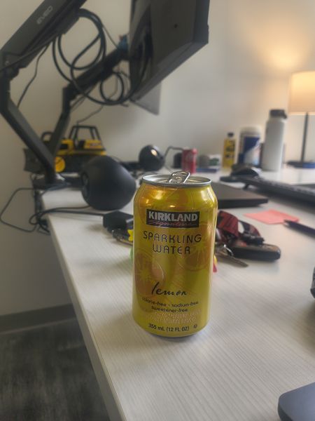 The image shows a can of Kirkland Signature Lemon Sparkling Water placed on a desk in an office setting. The can is yellow with a black label and features a lemon illustration. The desk is cluttered with various items including a computer monitor, headphones, a lamp, and other office supplies.