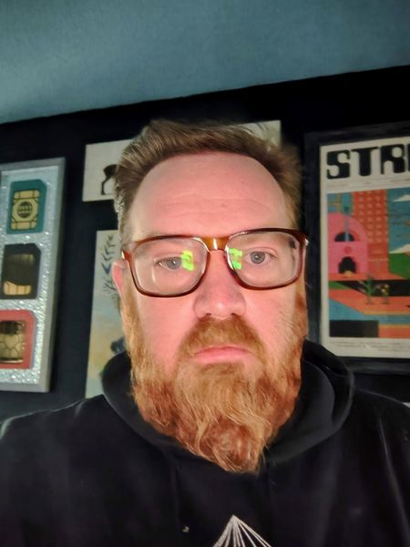 The image features a person with a reddish-brown beard and glasses, wearing a black hoodie with a white geometric design on the chest. The background includes framed posters and artwork, with one prominently displaying the word "STR" in bold letters. The setting appears to be indoors, with a dark blue wall and a black frame surrounding the artwork.