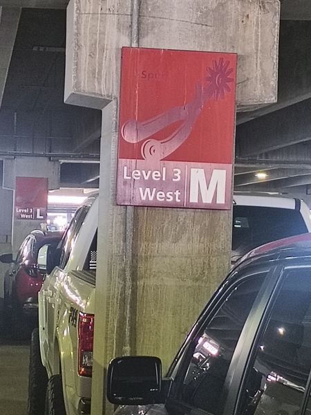 The image shows a parking garage with a prominent red sign labeled "Level 3 West" featuring a stylized cowboy boot graphic and the letter "M." The sign is mounted on a concrete pillar, and the setting includes parked vehicles, including a yellow Ford truck and a black car. The garage has a concrete ceiling with visible beams and lighting fixtures.