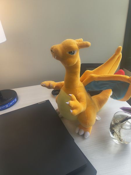 The image features a plush toy of a yellow dragon-like creature with blue wings, positioned on a desk. The setting includes a Lenovo laptop, a clear glass with a straw, and a black lamp with a blue base. The background is a plain beige wall.