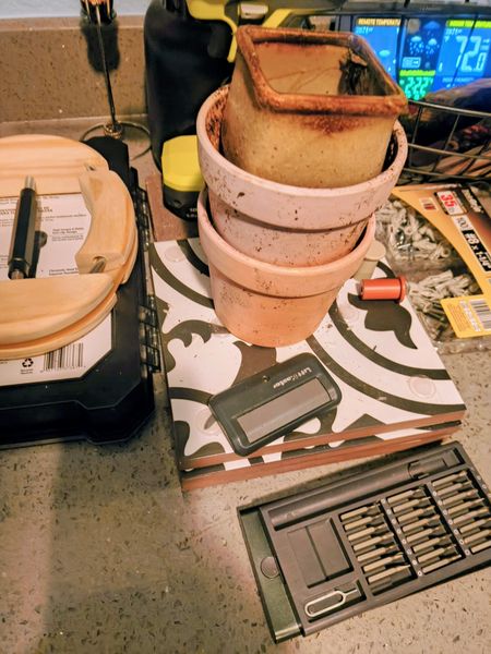 The image shows a stack of terracotta pots on a patterned cutting board, surrounded by various tools and hardware items, including a black and yellow cordless drill, a screwdriver set, and a package of screws. The setting appears to be a kitchen countertop, with a digital thermometer displaying a temperature of 72°F in the background.