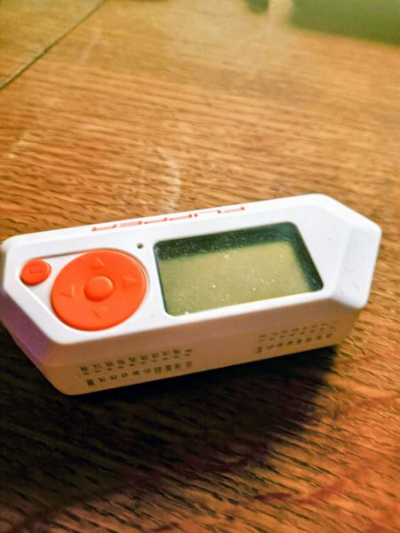 A white handheld electronic device with a green screen and orange buttons, possibly a game or toy, is placed on a wooden surface. The device features a prominent red circular button and some markings, though specific brand names are not clearly visible.