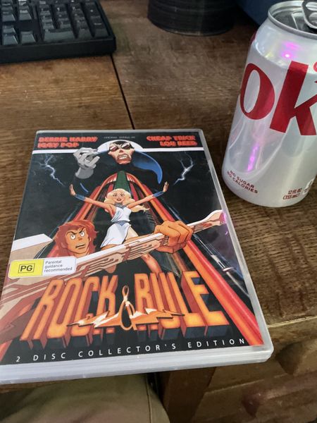 The image features a "Rock & Rule" DVD Collector's Edition placed on a wooden surface next to a can of Diet Coke. The DVD cover art includes a stylized illustration of a woman in a white dress, surrounded by dynamic, electric-themed artwork. The setting appears to be a casual indoor environment, with a keyboard partially visible in the background.