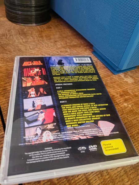 The back cover of a "Rock & Rule" DVD case lies on a light wooden table, displaying a vertical filmstrip of animated character stills on the left and yellow text listing special features for Disc 1 and Disc 2. Notable details include a yellow "Parental guidance recommended" label and a DVD Video logo in the bottom right, with a blue mesh object and a stack of dark discs visible in the background.