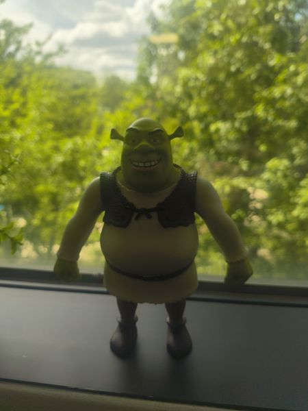 The image features a Shrek action figure standing on a windowsill, with a blurred background of lush green trees and a partly cloudy sky. The figure is dressed in a black vest over a yellow outfit and has a cheerful expression.