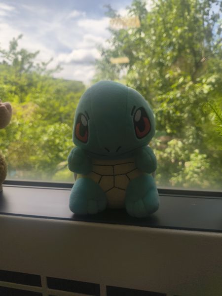The image features a plush toy of Squirtle, a green turtle Pokémon, sitting on a windowsill. The background is a blurred view of lush green trees and a partly cloudy sky, suggesting a peaceful outdoor setting. The Squirtle plush is light green with a yellow belly and red eyes, positioned in front of a window with a white frame.