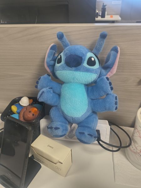 The image features a large, plush blue character, resembling Stitch from Disney's "Lilo & Stitch," sitting on a white power strip. The setting appears to be a desk or workspace, with a black smartphone, a white box labeled "Moo," and a white mug with red markings visible in the foreground. The background includes a wooden surface and part of a black object, possibly a speaker or electronic device.