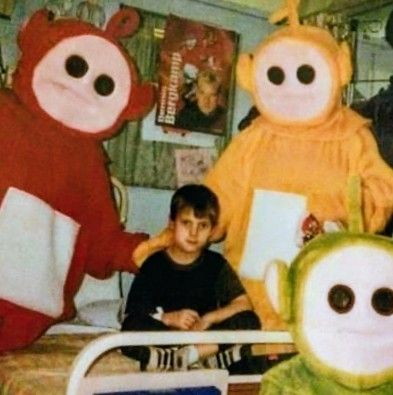 The image features a young boy sitting on a hospital bed, flanked by three Teletubbies characters in their iconic costumes: Dipsy in red, Laa-Laa in yellow, and Tinky Winky in green. The setting appears to be a hospital room, with posters and a curtain in the background. The boy is wearing a dark shirt and shorts, and the Teletubbies are holding papers, seemingly interacting with him.