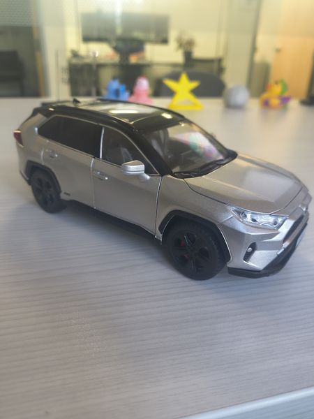 The image features a silver Toyota RAV4 model car with black wheels and silver accents, placed on a light gray textured surface. The background includes a glass partition, a yellow star-shaped object, and some colorful toys, suggesting a playful or casual setting.
