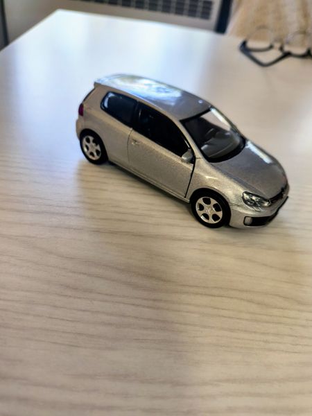 The image features a silver toy car, resembling a Volkswagen, positioned on a light wooden surface. The car has a shiny, metallic finish and is detailed with visible wheels and windows. The background includes a blurred view of a laptop and a pair of glasses, suggesting the setting is likely a desk or workspace.