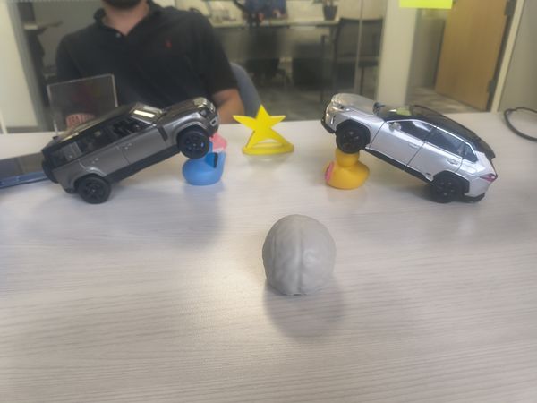 The image shows a white, brain-shaped object in the foreground on a light-colored table. In the background, two toy cars, one gray and one silver, are positioned on small yellow and blue bases, with a yellow star-shaped object between them. A man in a black polo shirt is partially visible, seated at a table in the background, which has a glass partition. The setting appears to be an office or meeting room.