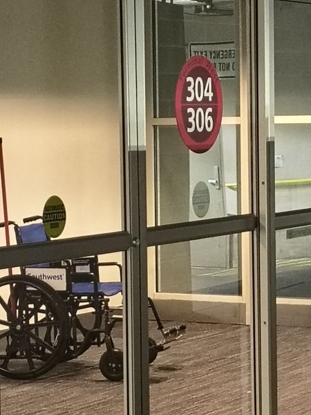 The image shows a glass door with a red circular sign displaying the numbers "304" and "306." Inside, a blue wheelchair with a "Southwest" sticker is visible, along with a small scooter. The setting appears to be a medical or institutional environment, indicated by the wheelchair and scooter, and the sign on the door suggests it is an emergency exit.