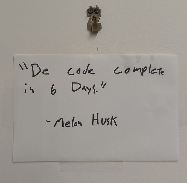 The image shows a handwritten note pinned to a white wall with a brass-colored hook. The note reads, "Be code complete in 6 Days." - Melon Husk. The handwriting is in black ink on a white piece of paper.