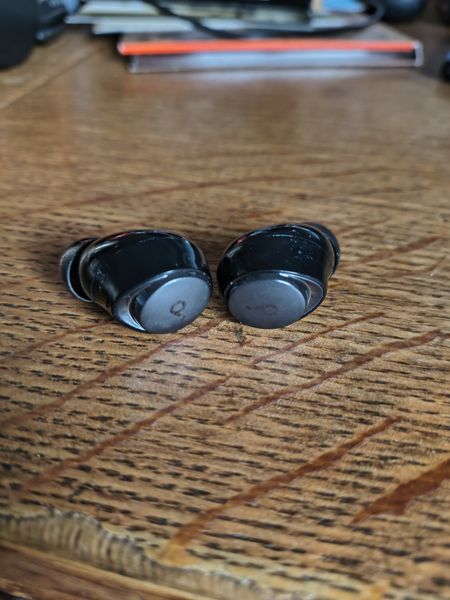 The image shows a pair of black wireless earbuds resting on a wooden surface with visible grain patterns. The background includes blurred objects, possibly books or papers, and a hint of a red pen or similar item.