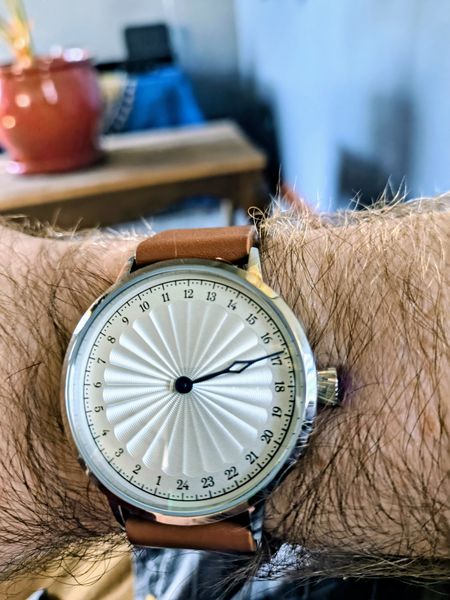 The image showcases a close-up of a wrist adorned with a watch featuring a white, textured dial with black hour markers and hands, set against a silver bezel. The watch has a brown leather strap and is worn on a hairy forearm. The background is softly blurred, revealing a red ceramic pot and a blue object on a wooden surface, suggesting an indoor setting.